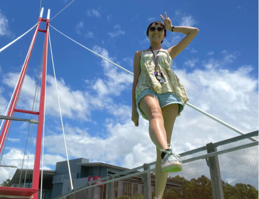 International student Jelisa stands on the Griffith University bridge showing a peace sign to the camera. Behind her the sky is blue and the weather is sunny